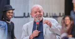 Lula veste terno cinza e camisa e segura microfone em uma palestra