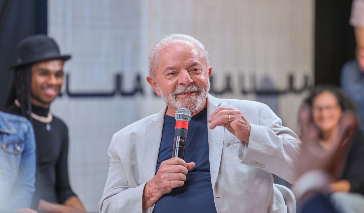 Lula veste terno cinza e camisa e segura microfone em uma palestra