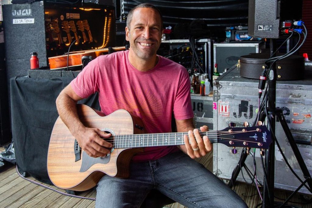Jack Johnson veste camisa de tom vermelho e segura um violão