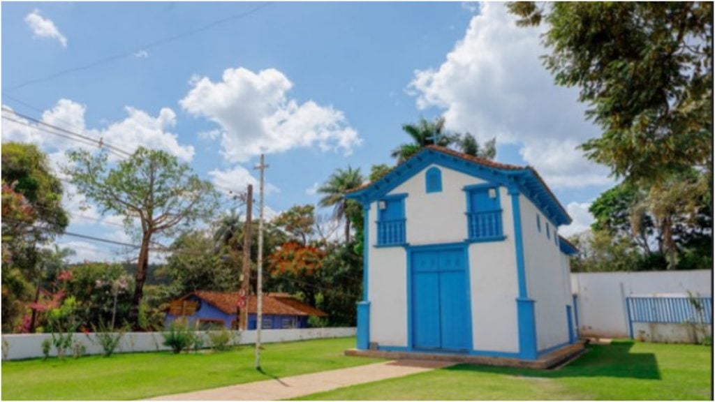 Capela azul e branca, com jardim verde, no centro de Macacos MG