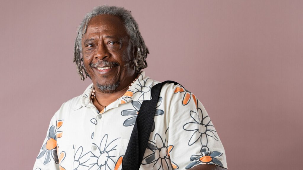 Homem negro, com cabelos grisalhos até a metade do rosto. Está sorrindo mostrando os dentes. Usa blusa branca com flores desenhadas em preto e laranja.