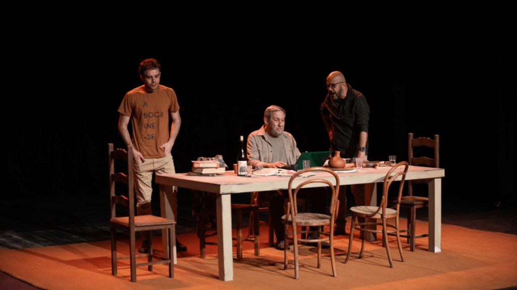 A imagem é de uma cena de teatro. Três homens estão reunidos em torno de uma mesa rústica, de cor clara, com cadeiras de madeira em um palco escuro, sob uma iluminação amarelada que foca no grupo. Ao centro, um homem mais velho, de barba e cabelo grisalhos, usa óculos e está sentado com um notebook na mesa, aparentemente digitando ou observando a tela. À sua esquerda, um homem jovem com uma camiseta marrom-laranja com os dizeres "A BOCA ME DINE-SE" (que pode ser uma frase em outro idioma) está em pé, com as mãos no bolso, olhando para o centro da cena. À direita, outro homem, careca e de barba escura, inclina-se sobre a mesa, gesticulando em direção ao notebook. A mesa está cheia de objetos, incluindo livros, uma garrafa escura e xícaras. O fundo é completamente preto e as cadeiras vazias em volta da mesa sugerem um cenário de reunião ou ensaio. A atmosfera é de concentração e colaboração.