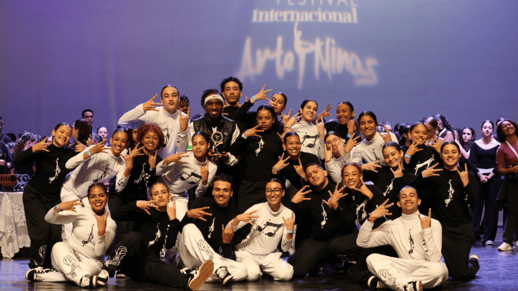 A imagem mostra um grande grupo de jovens dançarinos posando para uma foto de palco. A maioria das pessoas está fazendo gestos com as mãos, como chifres de roque ou o símbolo de paz, e há um sorriso generalizado. Eles estão vestindo roupas de dança em preto e branco, com designs que incluem silhuetas de notas musicais. O centro da imagem é dominado por um homem com uma bandagem na cabeça, um sorriso largo e um colar, que está no meio do grupo. O palco, o fundo e o chão estão iluminados, criando uma sensação de realização e celebração.