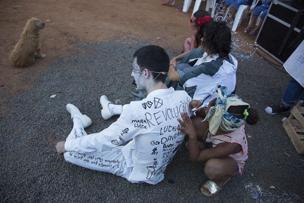 Homem participa do espetáculo sentado no chão, vestido com um terno branco, enquanto crianças escrevem sobre a roupa.